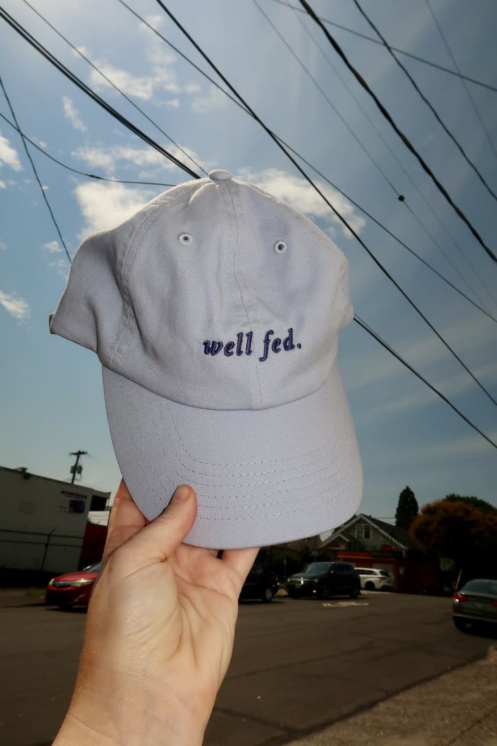 Blue Well-Fed Woman Hat held up outdoors against a blue sky, symbolizing empowerment and nourishment.