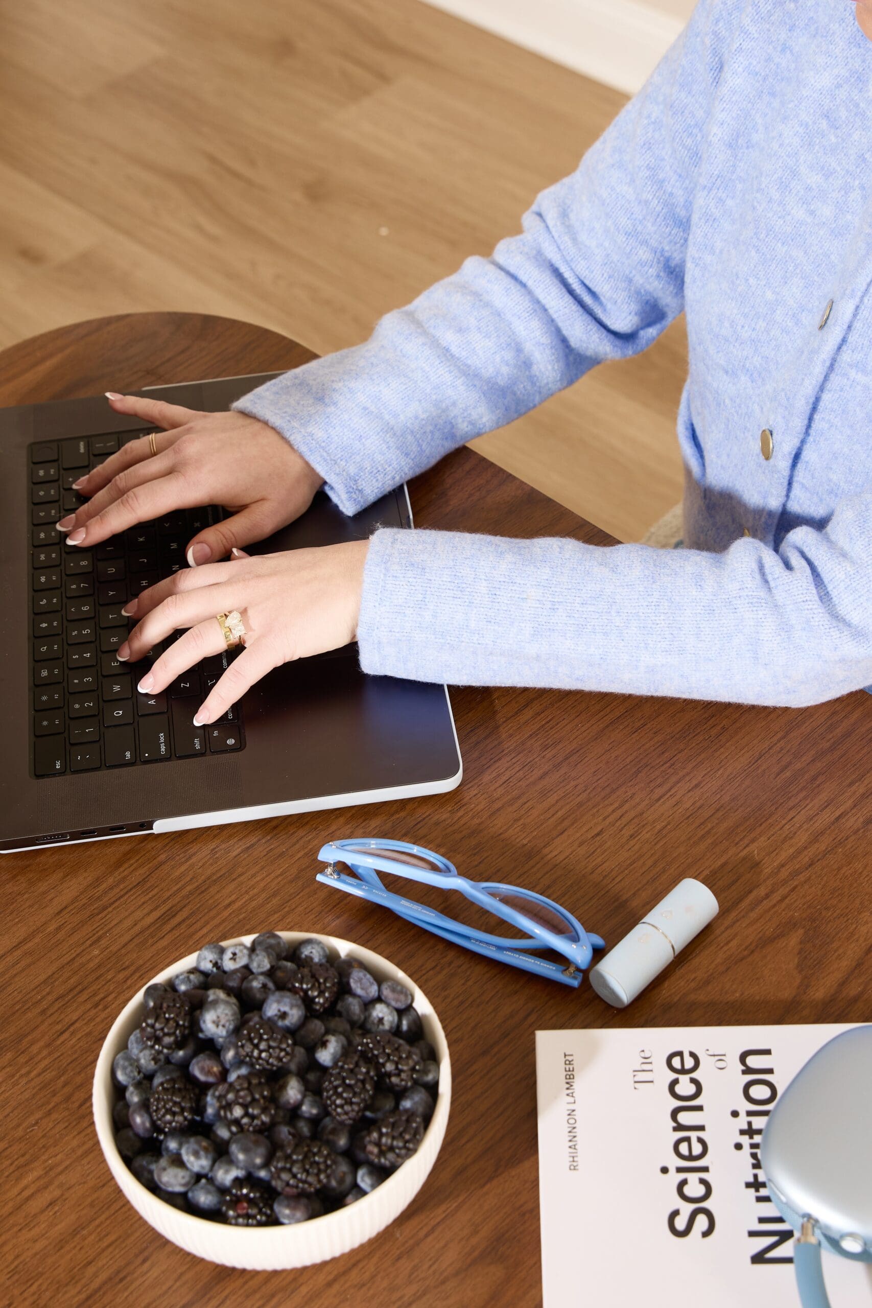 Registered dietitian working on a weekly meal plan at a laptop while snacking on fresh blackberries and blueberries, with blue headphones and glasses on the desk.