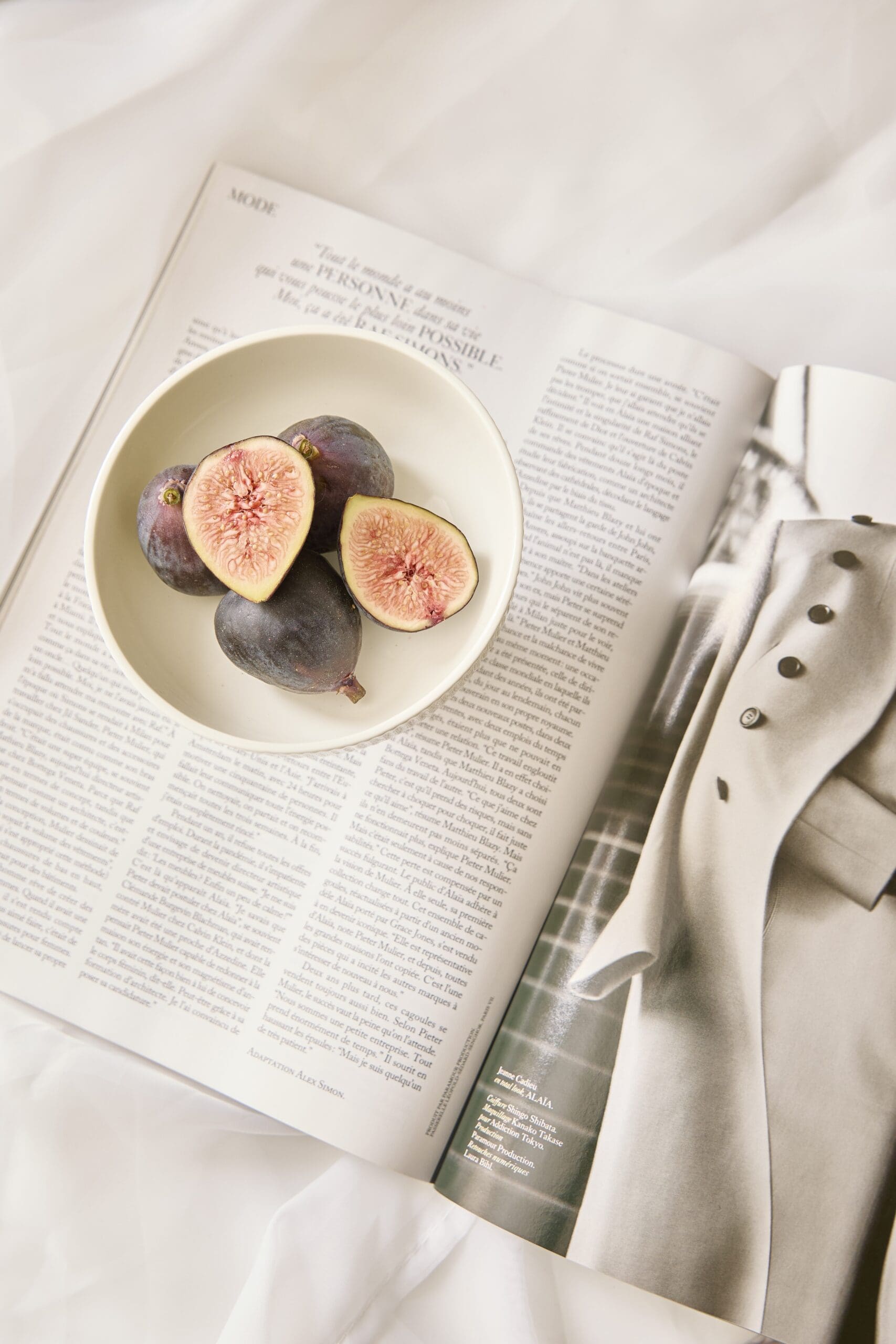 Bowl of fresh figs on an open magazine with a beige coat beside it.