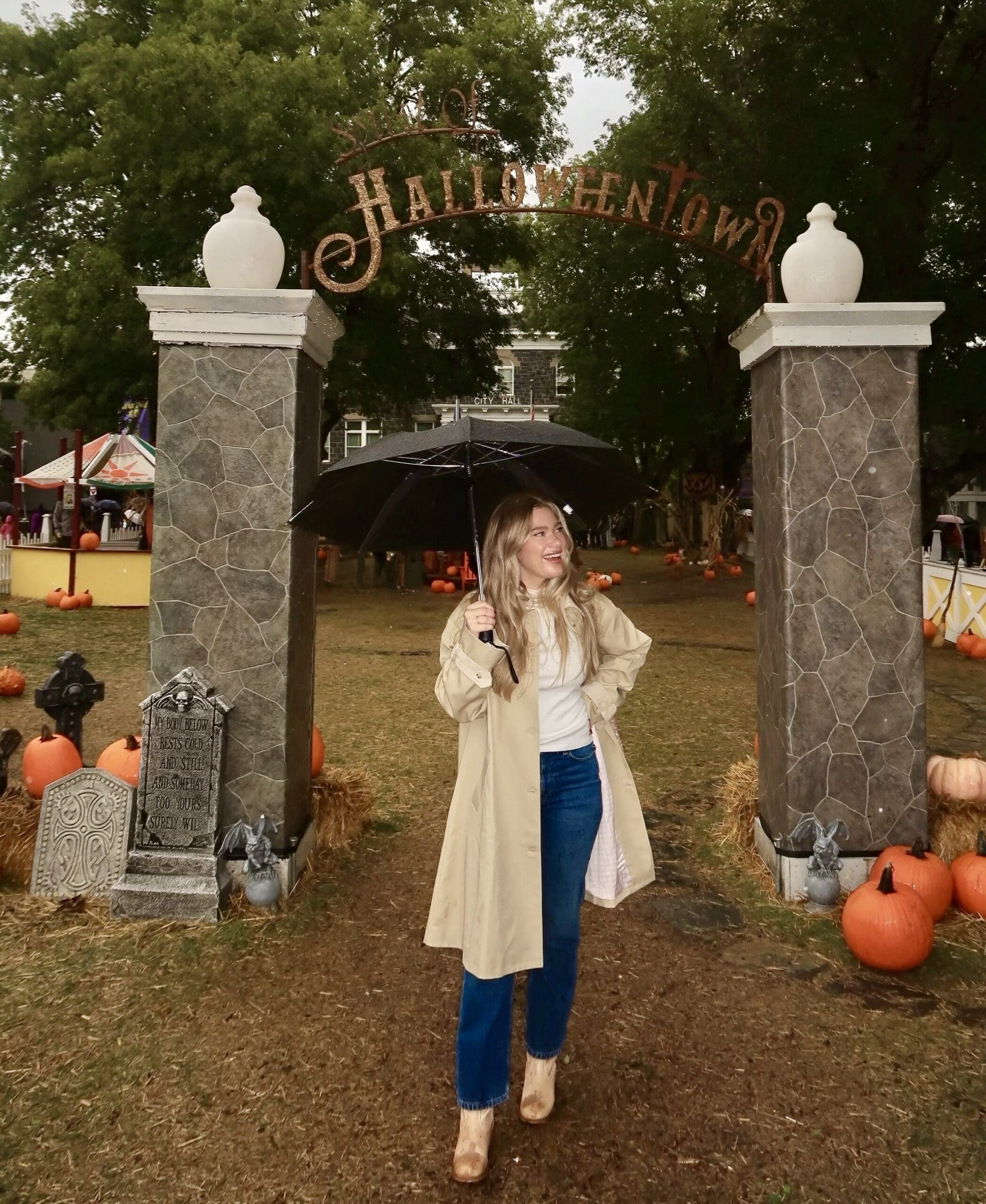 Claire Rifkin standing under the Halloweentown arch in Oregon on a rainy fall day, holding an umbrella and smiling in a beige trench coat and jeans surrounded by pumpkins.