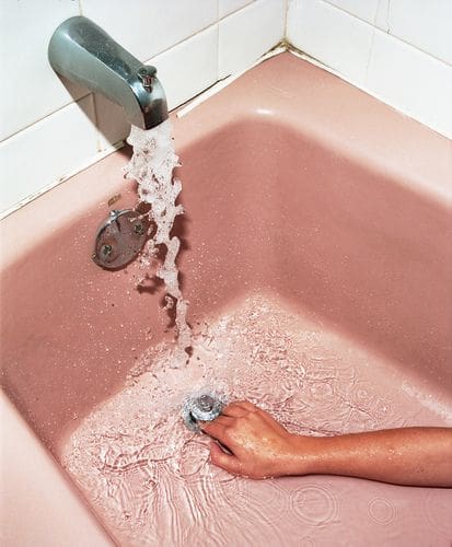 Hand cupping water beneath a running faucet in a pink bathtub, illustrating the struggle to hold water without a container.
